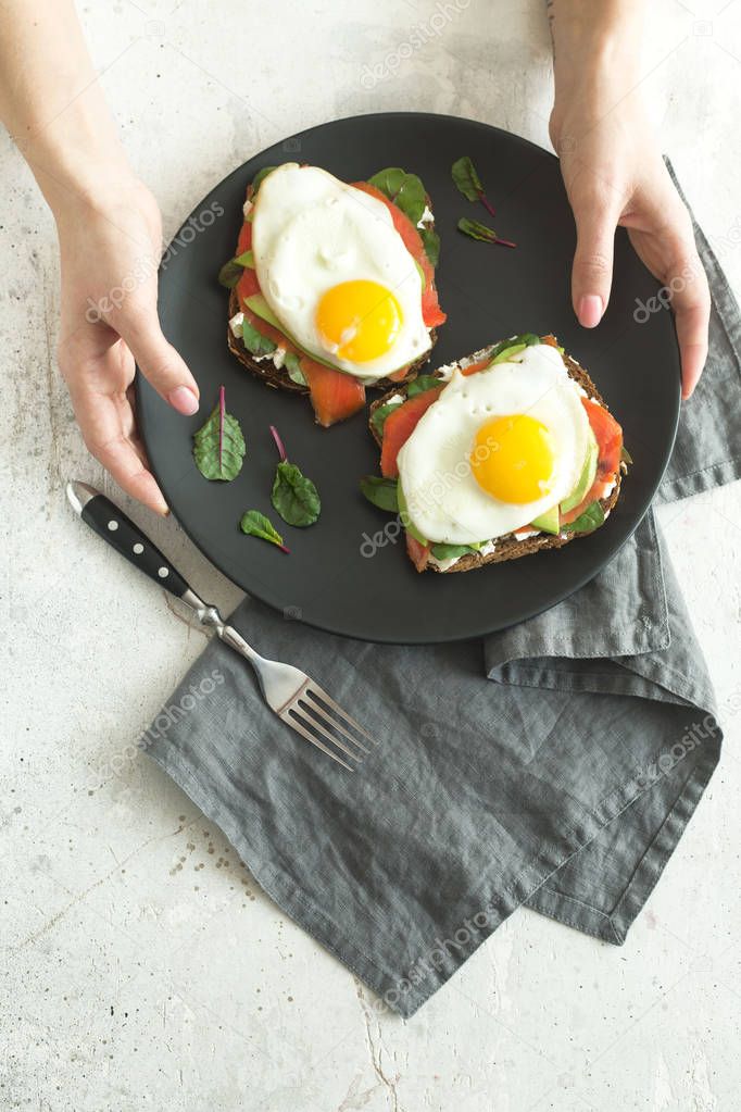Healthy breakfast sandwiches. Woman holding plate with bread toasts with fried eggs and fresh vegetablesover grey marble background, top view. Clean eating, healthy, diet, weight loss food concept