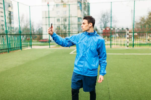 The guy is listening makes a selfie during a workout. A young man plays sports, runs on the football field. The guy works in the open, fresh air.