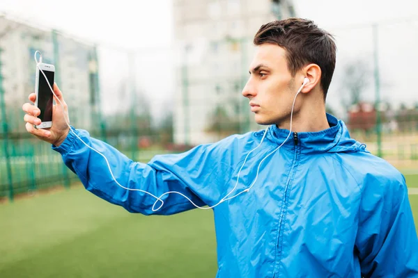 The guy is listening makes a selfie during a workout. A young man plays sports, runs on the football field. The guy works in the open, fresh air.