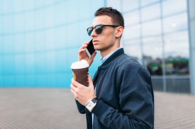 bir adam bir kağıt kahve, goes-den geçerek şehir, şık giysiler ve güneş gözlüğü, yakışıklı bir adam yapma bir telefon görüşmesi, iyi bir hava