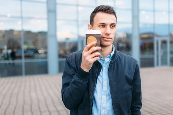 a man walks around the city and drinking coffee from a paper Cup, a handsome guy walks around and resting, the guy on the background of a modern building, office