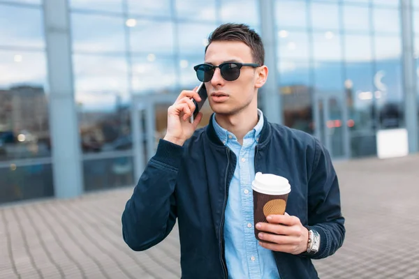 a man with a paper Cup of coffee, goes through the city, a handsome guy in stylish clothes and sunglasses, making a phone call, good weather