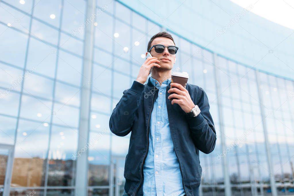 a man with a paper Cup of coffee, goes through the city, a handsome guy in stylish clothes and sunglasses, making a phone call, good weather