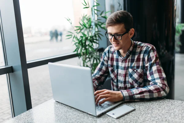 Un hombre se sienta en un café y trabaja en una computadora portátil, buscando trabajo, el estudiante escribe la tarea, un tipo con camisa y gafas — Foto de Stock