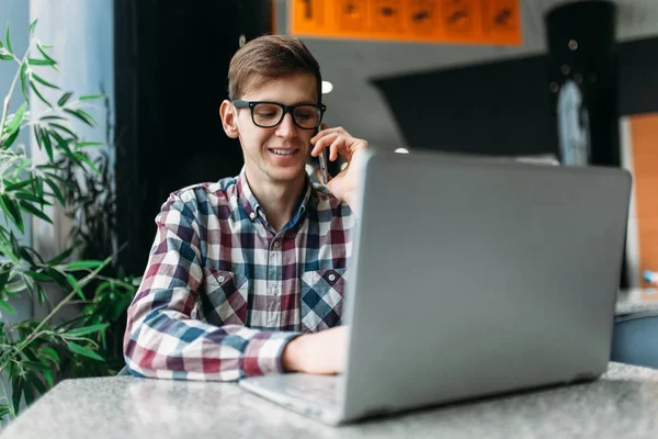 Un hombre se sienta en un café y trabaja en una computadora portátil, buscando trabajo, el estudiante escribe la tarea, un tipo con camisa y gafas — Foto de Stock