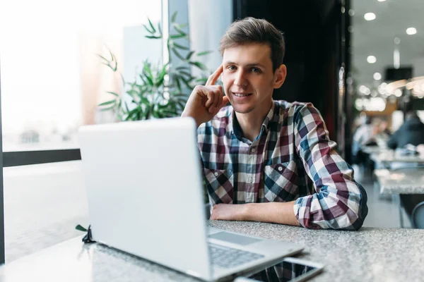 Un hombre se sienta en un café y trabaja en una computadora portátil, buscando trabajo, el estudiante escribe la tarea, un tipo con camisa y gafas — Foto de Stock