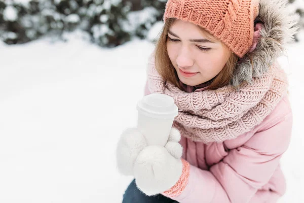 Retrato de uma bela jovem em branco mitenes de malha, ao ar livre, segurando um copo de papel com uma bebida quente, brilhante dia de inverno.A mulher sorri e é feliz, close-up, Árvores na primeira neve no . — Fotografia de Stock