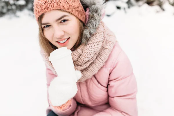 Портрет красивой молодой девушки в белых вязаных варежках, на открытом воздухе, проведение бумажной чашки с горячим напитком, яркий зимний день. Женщина улыбается и счастлива, крупным планом, деревья в первом снегу в . — стоковое фото
