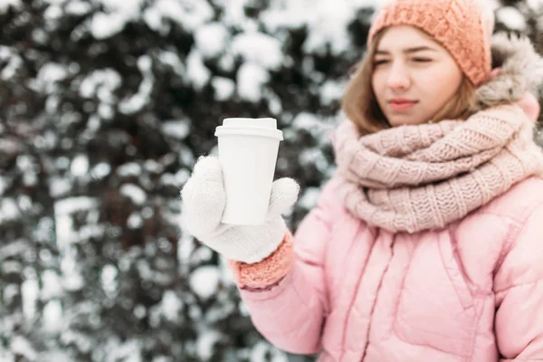 Retrato de uma bela jovem em branco mitenes de malha, ao ar livre, segurando um copo de papel com uma bebida quente, brilhante dia de inverno.A mulher sorri e é feliz, close-up, Árvores na primeira neve no . — Fotografia de Stock