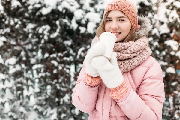 Retrato de uma menina bonita em mitenes de malha branca, ao ar livre, bebendo uma bebida quente, dia de inverno brilhante.mulher sorrindo e feliz, macro, árvores, primeira neve . — Fotografia de Stock