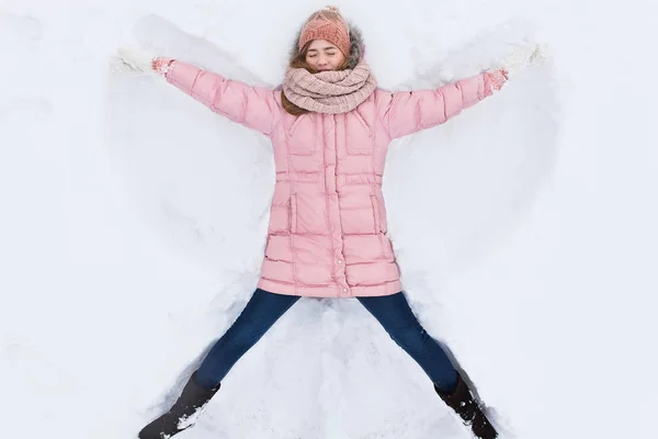 Счастливая женщина лежит в снегу и двигает руками и ногами вверх и вниз, создавая форму снежного ангела. Улыбающаяся женщина лежит на снегу в зимний отпуск, для рекламы  , — стоковое фото