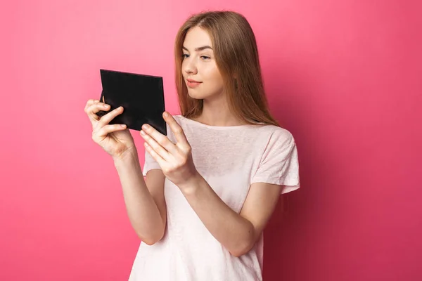 Retrato de una hermosa chica mirándose en el espejo, admirándose a sí misma, aislada sobre fondo rosa, feliz y linda — Foto de Stock