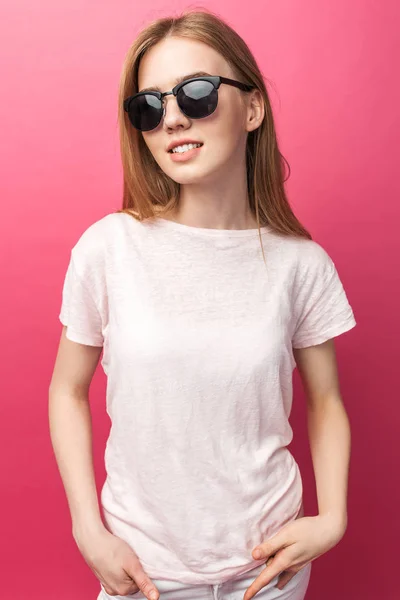 Hermosa joven rubia en gafas de pie sobre fondo rosa con pantalones vaqueros, top rosa sonrisa blanca nieve sonriente, con gafas negras y buscando la foto promocional perfecta —  Fotos de Stock