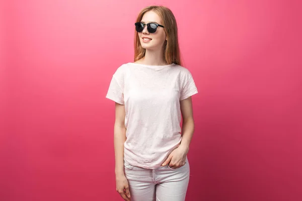 Menina loira bonita em óculos de pé sobre fundo rosa vestindo jeans, rosa top sorrindo neve sorriso branco, vestindo óculos pretos e olhando foto promocional perfeita — Fotografia de Stock