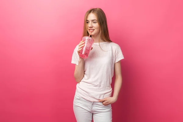 Retrato de bela jovem bebendo bebida gasosa de uma garrafa com uma palha e olhando para a câmera isolada em um fundo rosa, é o texto — Fotografia de Stock