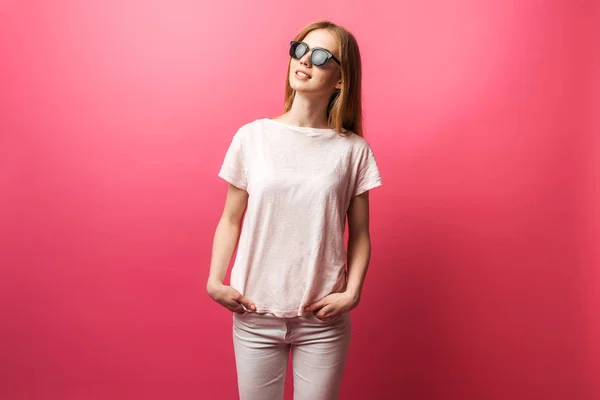 Bella giovane ragazza bionda in occhiali in piedi su sfondo rosa indossando jeans, top rosa sorriso bianco neve, indossando occhiali neri e cercando perfetta foto promozionale — Foto Stock