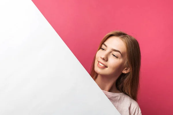 Όμορφη νεαρή κοπέλα που κρυφοκοιτάζουν έξω από πίσω από λευκό χαρτί τοίχο, σε ροζ φόντο — Φωτογραφία Αρχείου