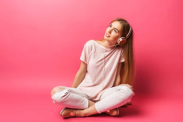 Retrato de alegre chica hermosa sentada en el suelo y mirando hacia otro lado, escuchando música en auriculares, aislada sobre fondo rosa, publicidad — Foto de Stock