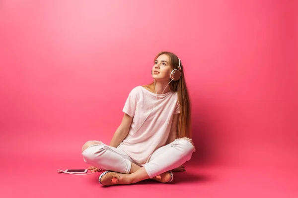 바닥에 앉아 멀리 보고, 이어폰에 고립 된 분홍색 배경, 광고 음악을 듣고 밝은 아름 다운 여자의 초상화 — 스톡 사진