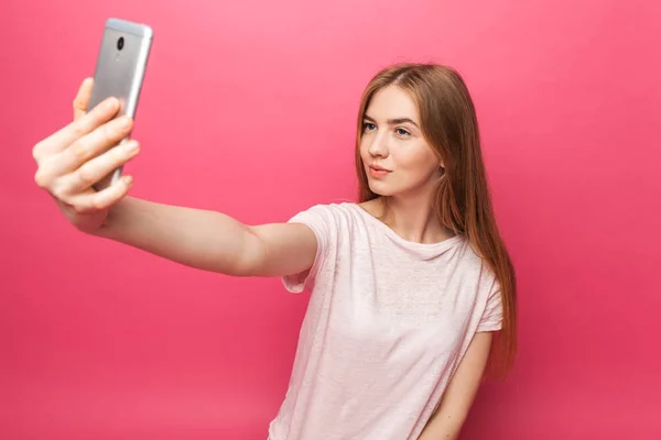 Retrato de chica hermosa alegre, fotografiado, tomando selfie, mirando en el teléfono, bastante divertido, aislado en el fondo rosa, publicidad — Foto de Stock