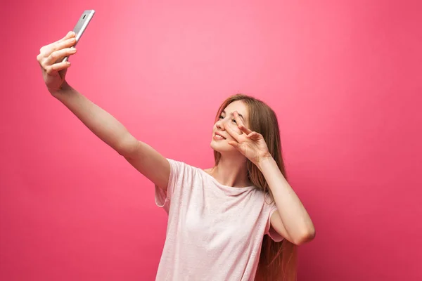 Retrato de chica hermosa alegre, fotografiado, tomando selfie, mirando en el teléfono, bastante divertido, aislado en el fondo rosa, publicidad — Foto de Stock