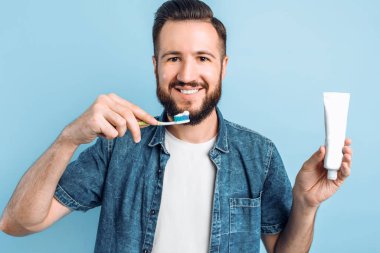 Sakallı, genç ve yakışıklı bir adam dişlerini kaldırılmış mavi bir arka planda fırçalıyor.