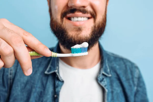 A young handsome man with a beard is brushing his teeth on an isolated blue background with raised — Stock Photo, Image