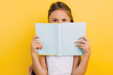 Smart teen girl looks out of a book and looks at the camera isolated on a yellow background