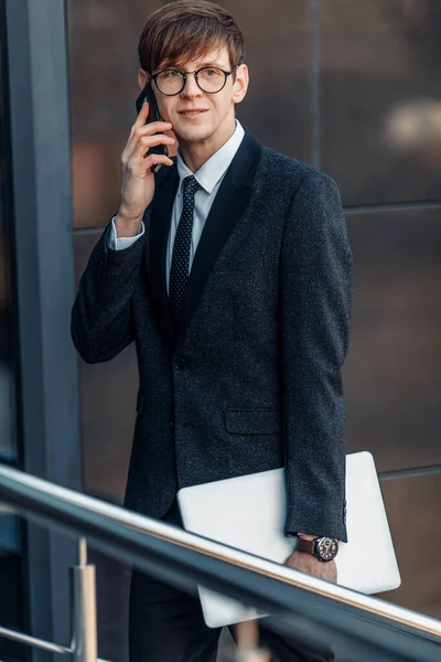 Confident man-businessman in a business suit, talking on the phone in the open air with an office building in the background