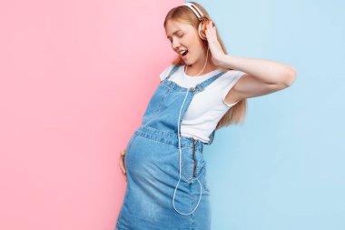 Mutlu şirin, güzel, neşeli, hamile bir kadın, izole edilmiş pembe ve mavi bir arka planda dururken kulaklıkla müzik dinlemekten zevk alıyor.