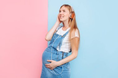 Mutlu şirin, güzel, neşeli, hamile bir kadın, izole edilmiş pembe ve mavi bir arka planda dururken kulaklıkla müzik dinlemekten zevk alıyor.