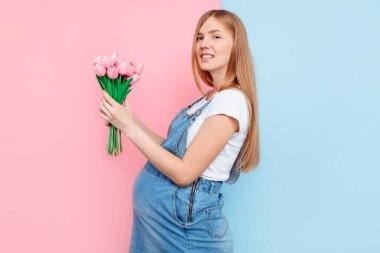 Pembe ve mavi bir arka planda duran bir buket lale ile mutlu, hamile bir kadın. Anneler Günü konsepti