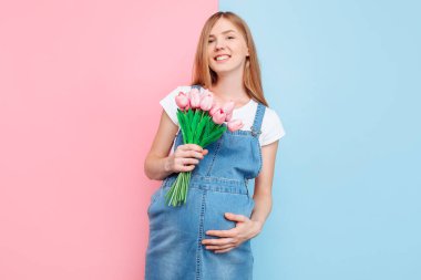 Pembe ve mavi bir arka planda duran bir buket lale ile mutlu, hamile bir kadın. Anneler Günü konsepti