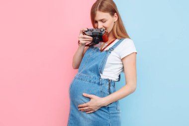 Kot elbiseli hamile güzel bir kadın pembe ve mavi arka planda izole edilmiş eski bir kamerada fotoğraf çekiyor.