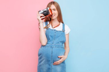 Kot elbiseli hamile güzel bir kadın pembe ve mavi arka planda izole edilmiş eski bir kamerada fotoğraf çekiyor.