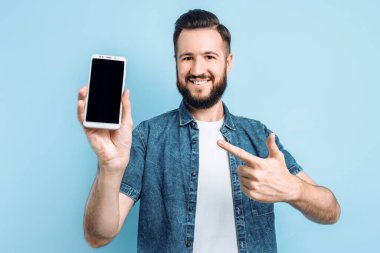 Elinde akıllı bir telefon tutan ve kameraya boş bir akıllı telefon ekranı gösteren mutlu genç bir adam mavi bir torba üzerinde onu parmağıyla işaret ediyor.