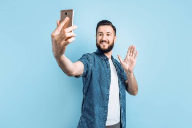 Sıradan giysiler içinde sakallı, mutlu modaya uygun bir adam, gülümseyen ve akıllı telefondan selfie çeken, mavi bir arka planda duruyor.