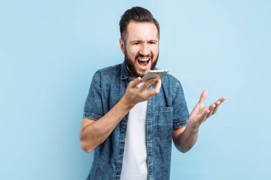 Heyecan verici bir şekilde bağıran bir adam telefonuyla konuşuyor ve telefona bağırıyor, mavi bir arka planda duruyor.