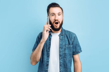 Şok olmuş sakallı genç bir adam telefonda konuşuyor ağzı açık, şaşırmış bir şekilde, izole edilmiş mavi bir arka planda duruyor.