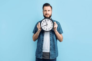 Sakallı düşünceli genç bir adam, izole edilmiş mavi bir arka planda duran duvar saatini tutuyor.