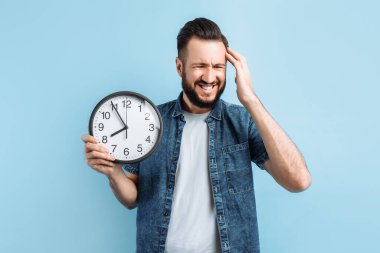 Bitkin, geç gelen sakallı genç bir adam, izole edilmiş mavi bir arka planda duran bir duvar saati tutuyor.