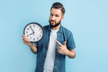 Mavi arka planda duvar saati tutan sakallı ve şok olmuş genç bir adam.