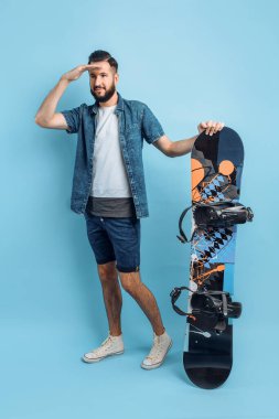 Şort ve gömlek giymiş mutlu sakallı bir adam, bir snowboard tutuyor ve uzaklara bakıyor, izole edilmiş mavi bir arka planda duruyor.