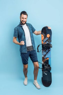 Sakallı, şort ve gömlek giyen mutlu bir adam, elinde bir snowboard tutuyor ve izole edilmiş mavi bir arka planda duran zafer ve başarının bir jestini sergiliyor.