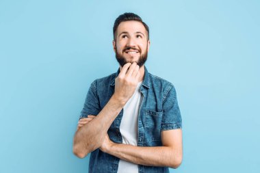 Akıllı ve düşünceli, sakallı, çenesini eliyle tutan, kaşlarını çatan ve üst köşeye bakan çekici bir adam mavi arka planda durarak bir karar verir.