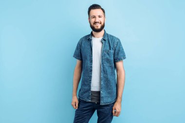Mutlu, modaya uygun, sakallı yakışıklı bir adam soyutlanmış mavi bir arka planda duruyor.