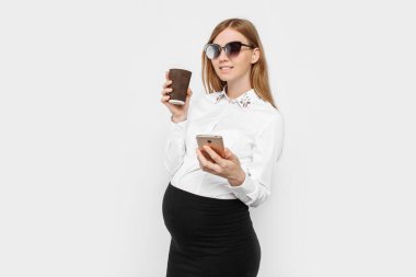 Gözlük takan genç hamile bir iş kadını, elinde akıllı bir telefon tutan ve bir fincan kahve içen bir kız, iş yerinde beyaz bir arka planda bir mola.