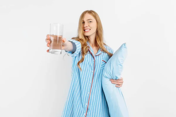 若いです美しい女性ですパジャマ姿で立って枕と白い背景に水のガラスを保持 — ストック写真