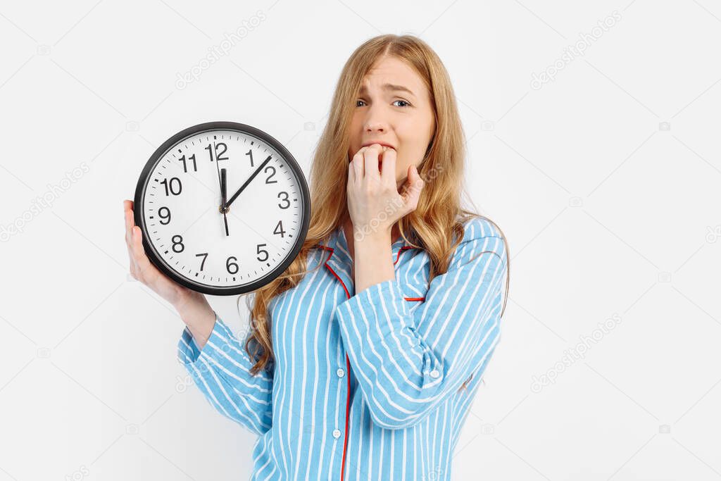 Image of a shocked and frightened girl in pajamas, with an alarm clock, holding her fingers near her mouth from fear, isolated on a white background