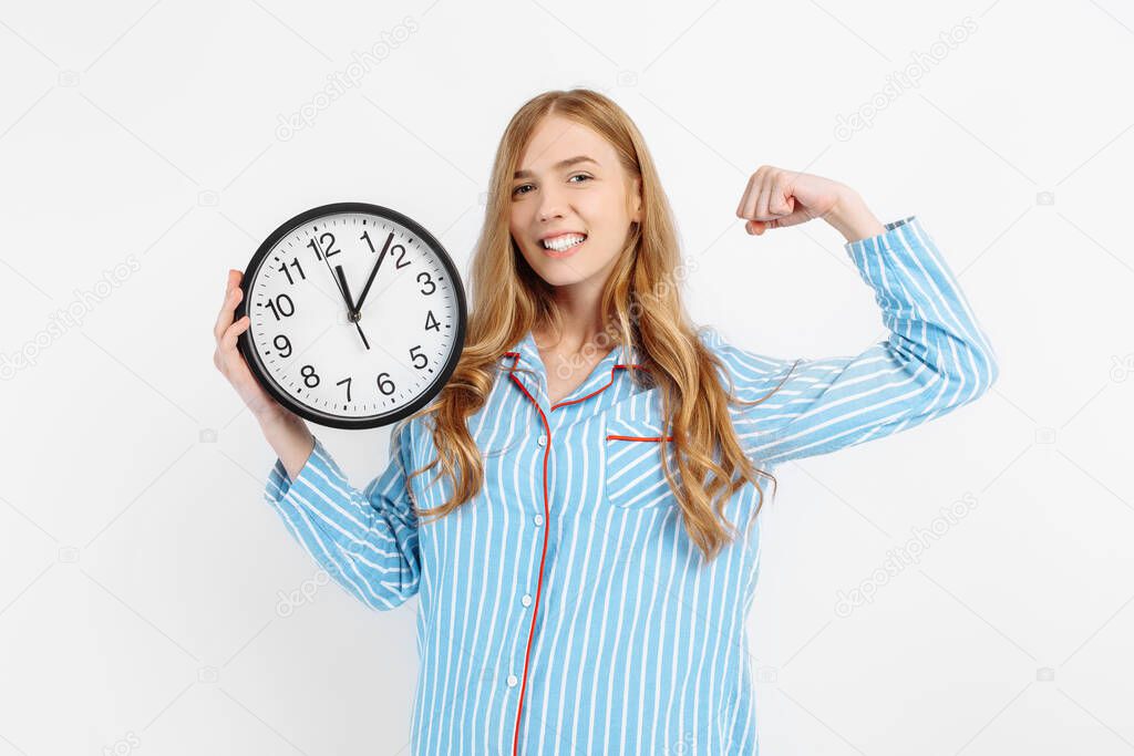 Beautiful girl in pajamas holding alarm clock and flexing biceps waking up early in the morning on white background